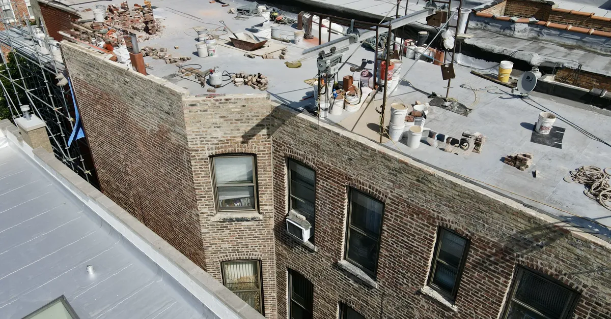 Drone aerial of a historic Chicago brick building with its parapet wall mid-rebuild: pallets of brick and repair rigging staged on the rooftop, original masonry still intact below